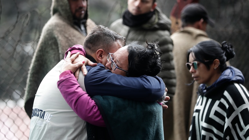 Two people hug next to a barbed wire fence as three others watch on. 