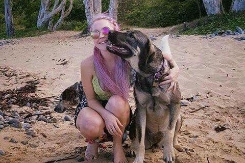 Toyah Cordingley, wearing pink sunglasses and with long pink hair, kneels on beach with her dog, date unknown.