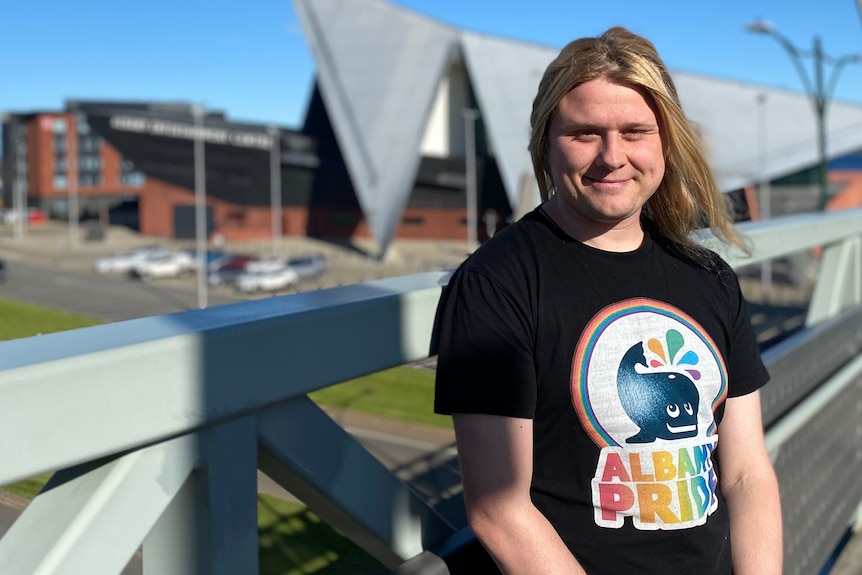 A smiling person with long blond hair wearing a black rainbow pride shirt stands on a bridge, buildings behind, blue sky.