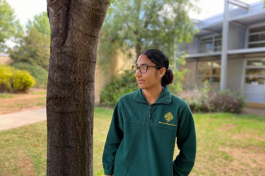 A woman is standing next to a tree, looking away from the camera.