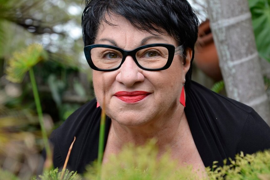 A woman with dark hair and dark rectangle glasses looks to the camera with a pursed smile. She has red lipstick on.