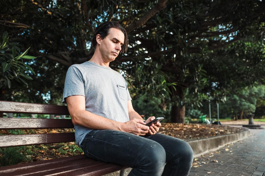 A man sitting on a wooden bench looking down at his mobile phone