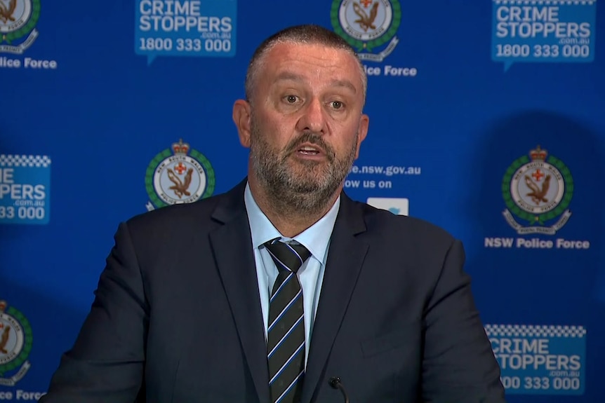 Detective Superintendent Joseph Doueihi stands in front of a wall with NSW Police badges and signs for crime stoppers