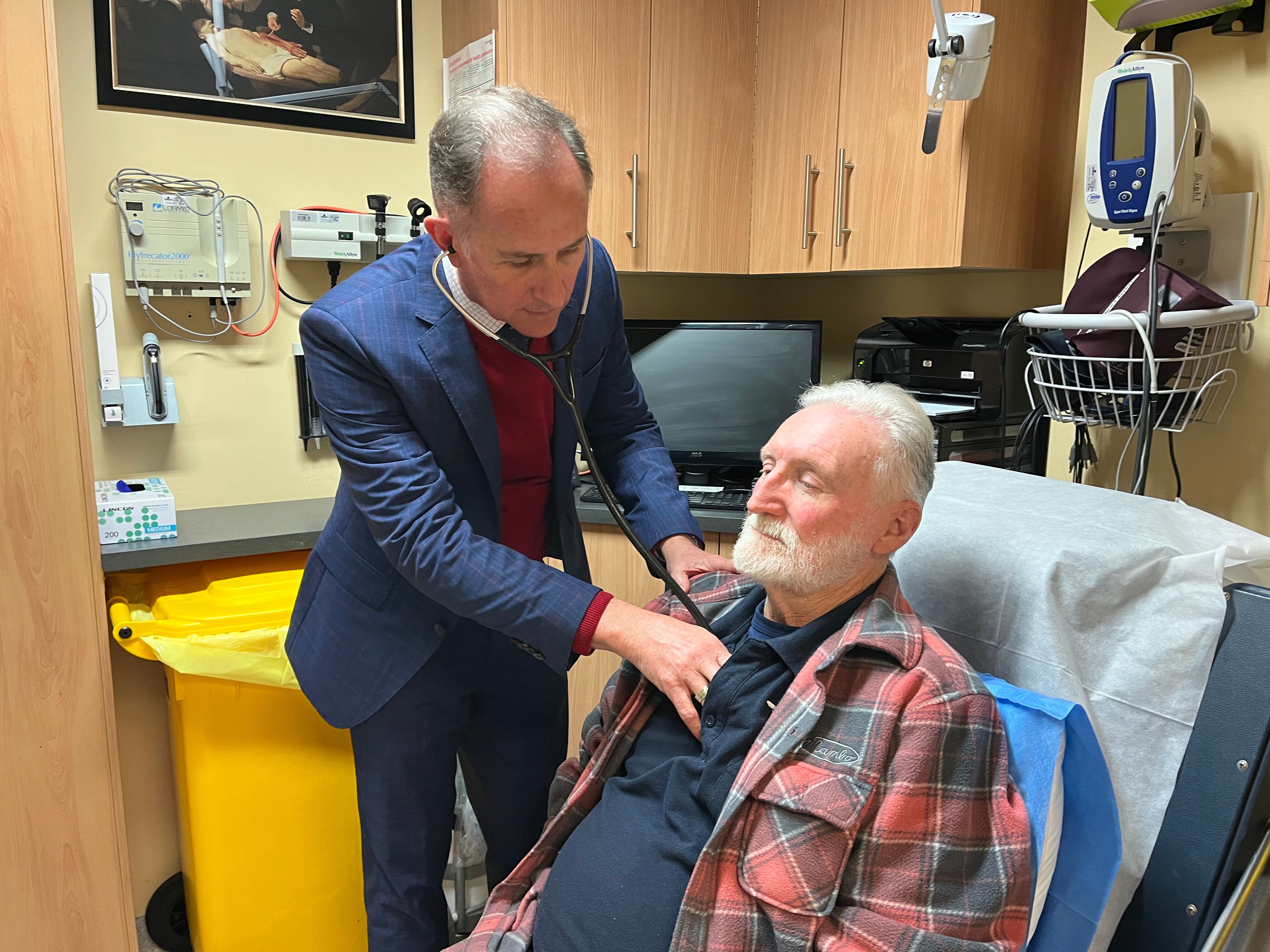 An older man wearing a suit, with a stethoscope around his neck, with an older male patient.