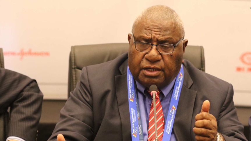 Wera Mori sitting at a desk speaking to delegates during an APEC meeting. He is wearing a suit and tie.