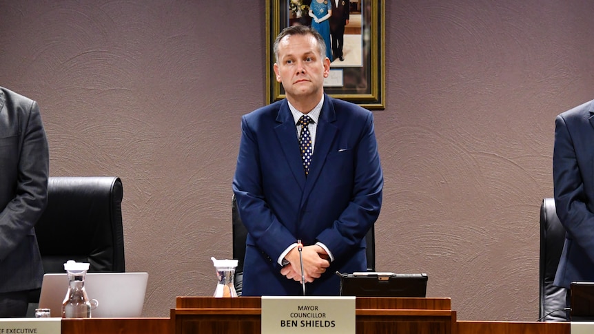 A man with short hair and a blue suit stands behind a desk