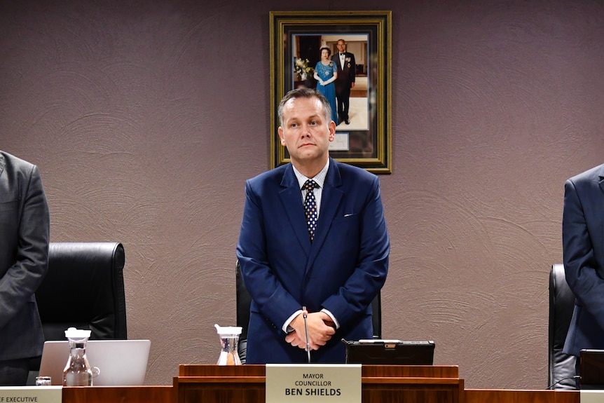 A man with short hair and a blue suit stands behind a desk