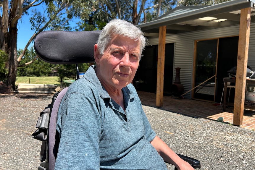 A middle aged white man with grey hair sitting in a wheelchair out the front of a farm house