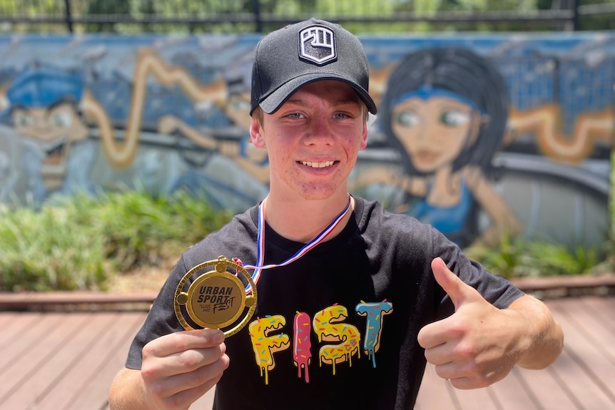 a teenager in a cap holds a gold coloured medal up to the camera and gives a thumbs up