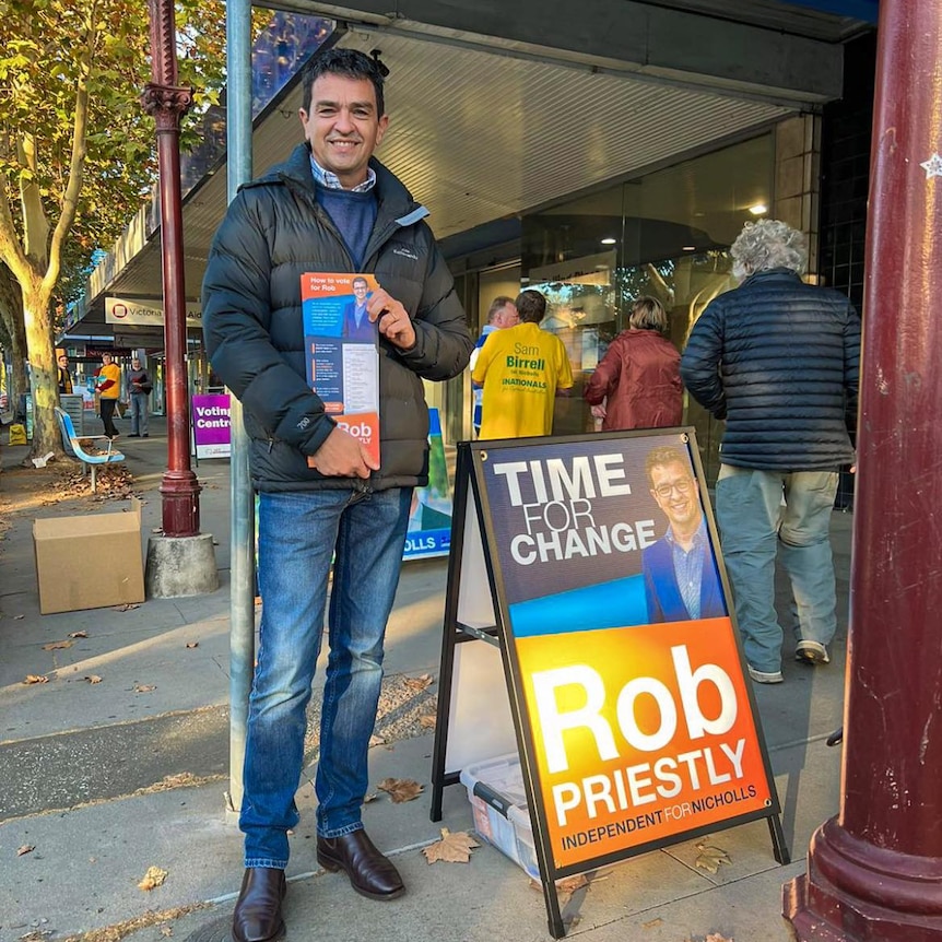 A dark-haired man in blue jeans and a jacket smiles while standing by one of his signs at an early voting centre