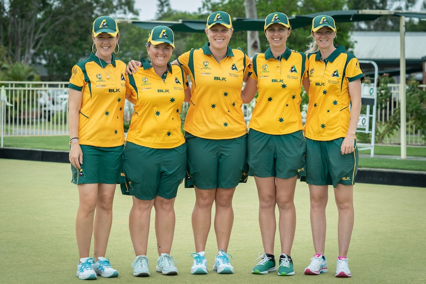 Five women in green and gold outfits stand on a bowling green, link arms and smile.