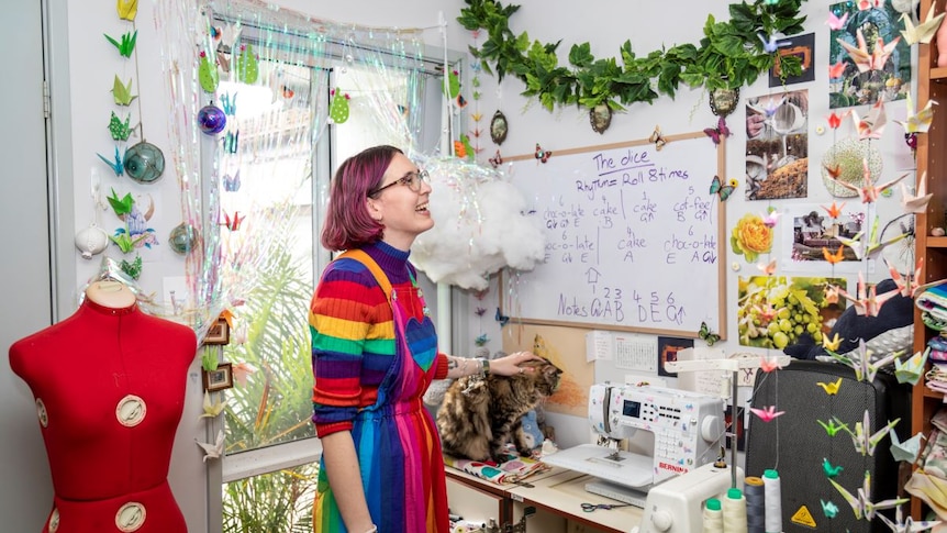 A young woman with pink hair, rainbow jumper and rainbow dress stands in a sewing room smiling.