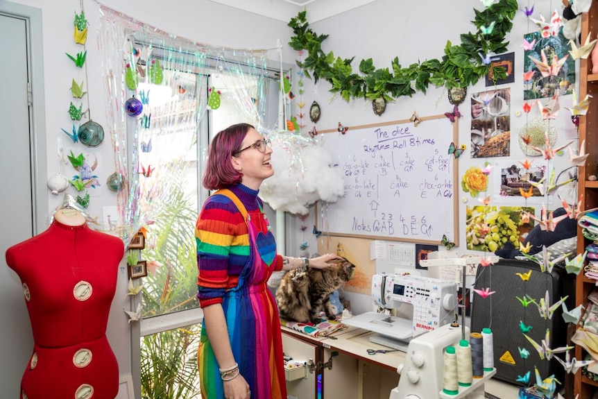 A young woman with pink hair, rainbow jumper and rainbow dress stands in a sewing room smiling.