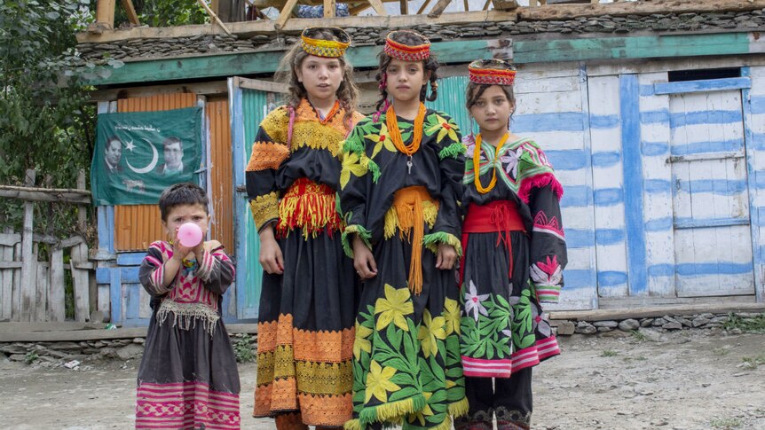 Four children in traditional costume stand by the side of a dirt road smiling at the camera