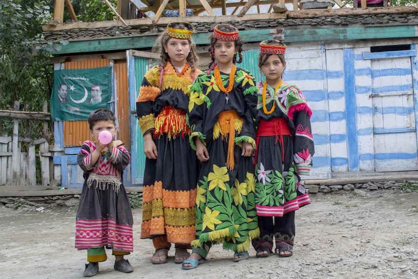 Four children in traditional costume stand by the side of a dirt road smiling at the camera
