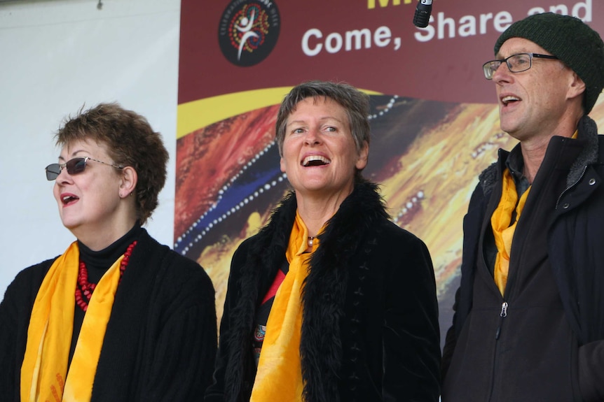A mid-shot of two women and a man stand on a stage singing wearing black coats and yellow scarves.