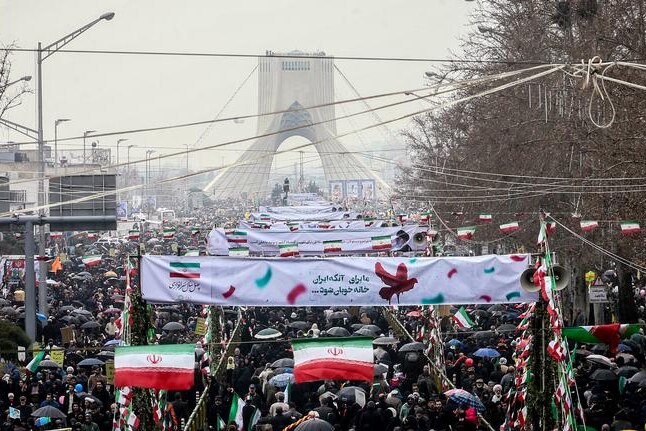 A sea of people spread across a Tehran street with placards an signs as a triangular monument sits on the horizon.