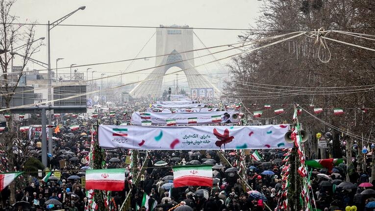 A sea of people spread across a Tehran street with placards an signs as a triangular monument sits on the horizon.