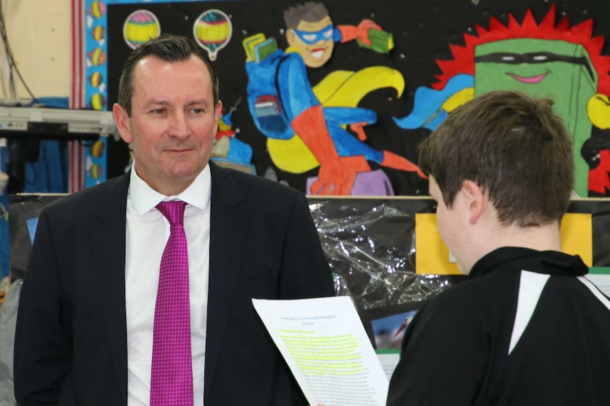 Premier Mark McGowan stands wearing a suit listening to a student read from a piece of paper at Kewdale Primary School.