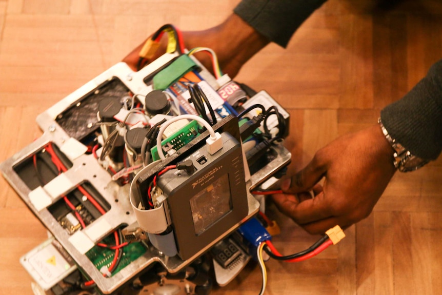 Close up of a robot with wires coming out of it as two hands plug red wires in