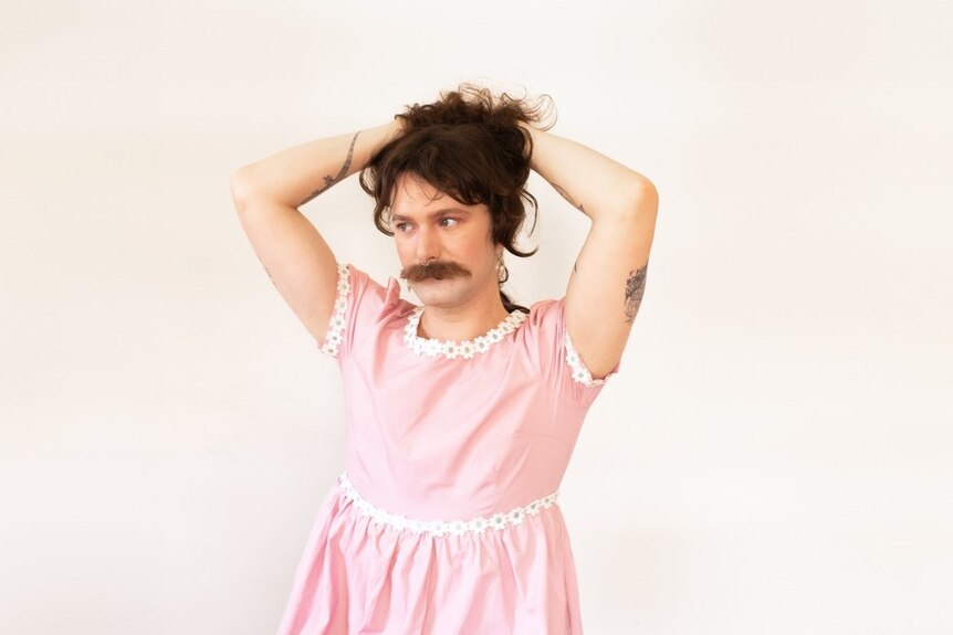 A person with long brown hair and a mustache dressed in a pink and floral dress  