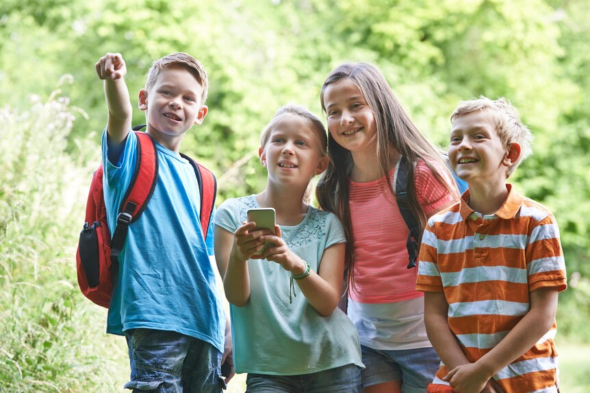 Four smiling school kids stand together in nature. One girl checks a smartphone and a boy points to the distance.