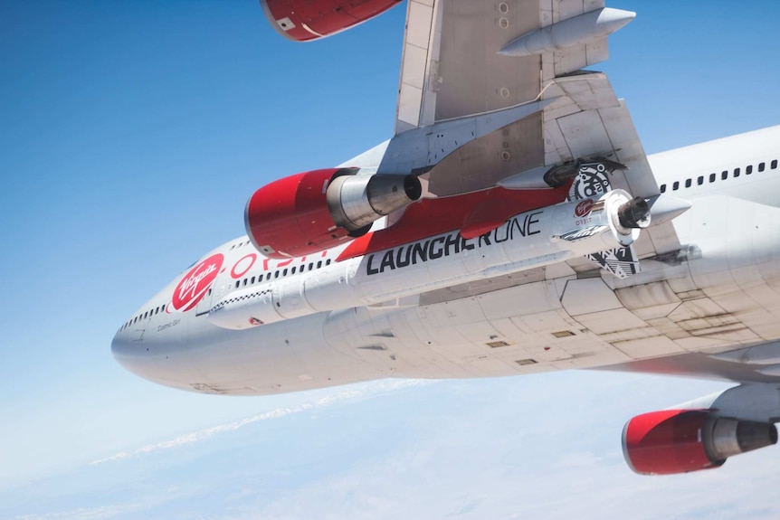 A rocket with Launcher One written on the side is attached underneath the wing of a Virgin branded 747.