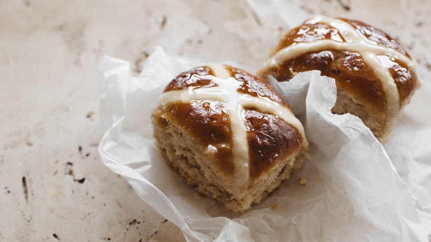 Two homemade hot cross buns on baking paper, an Easter baking recipe.