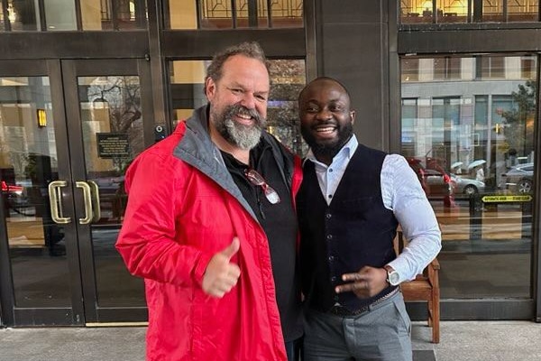 Two men stand next to each other, one in a red jacket and beard, the other in business attire, smiling 