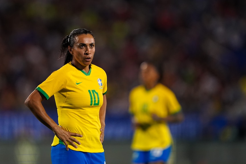 A Brazilian female international footballer stands with her hands on her hips during a match.