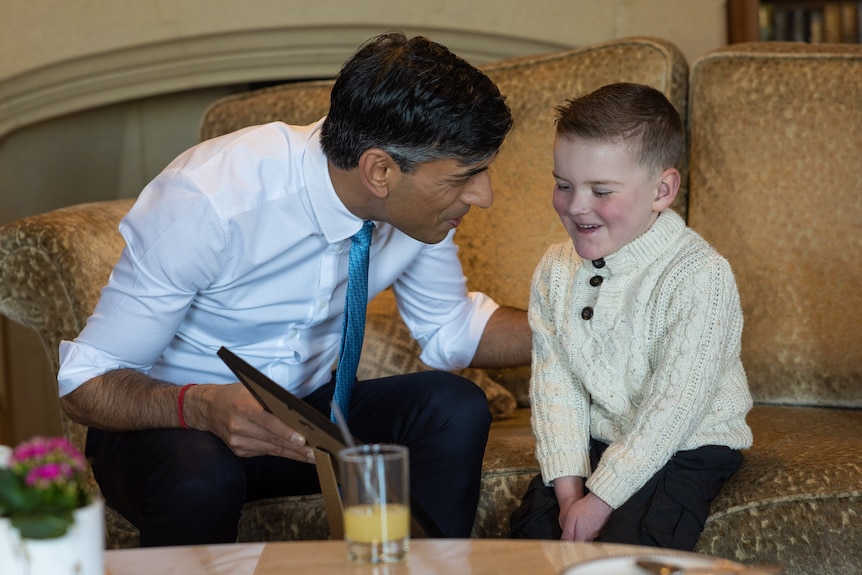 Rishi Sunak sits on a sofa with a young boy. Sunak is showing him a framed certificate, and the boy is grinning.