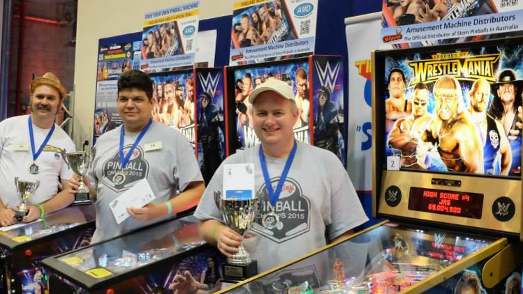 Three men standing in front of pinball machines holding trophies