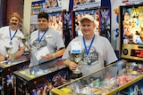 Three men standing in front of pinball machines holding trophies