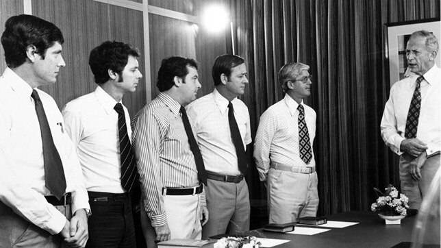 A black and white photograph of five ministers standing at a table.
