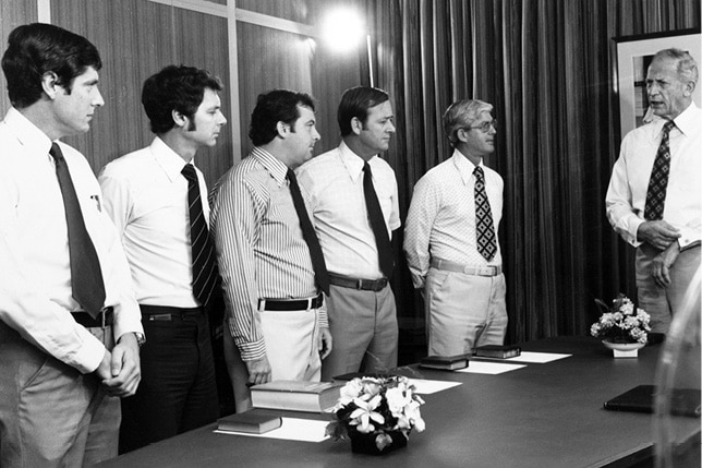 A black and white photograph of five ministers standing at a table.