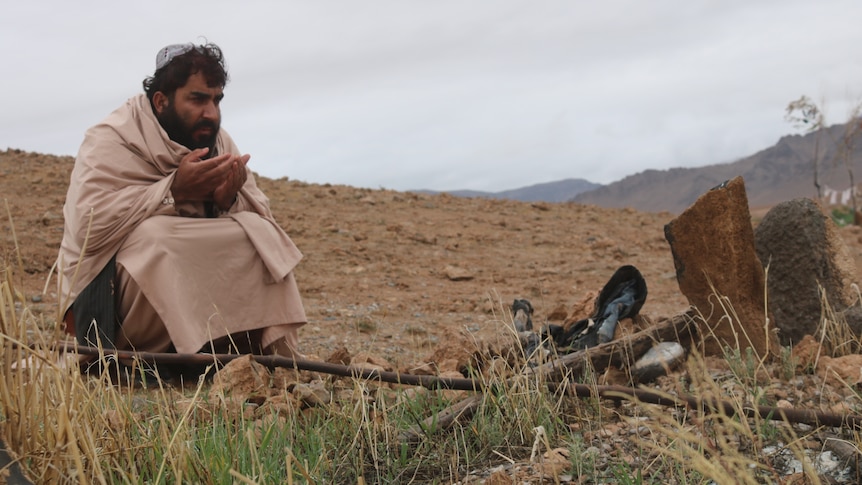 A man wrapped in a khaki shawl sits, praying beside a crude grave on a dusty mountain.