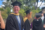 A man stands in a university graduation robe, smiling.