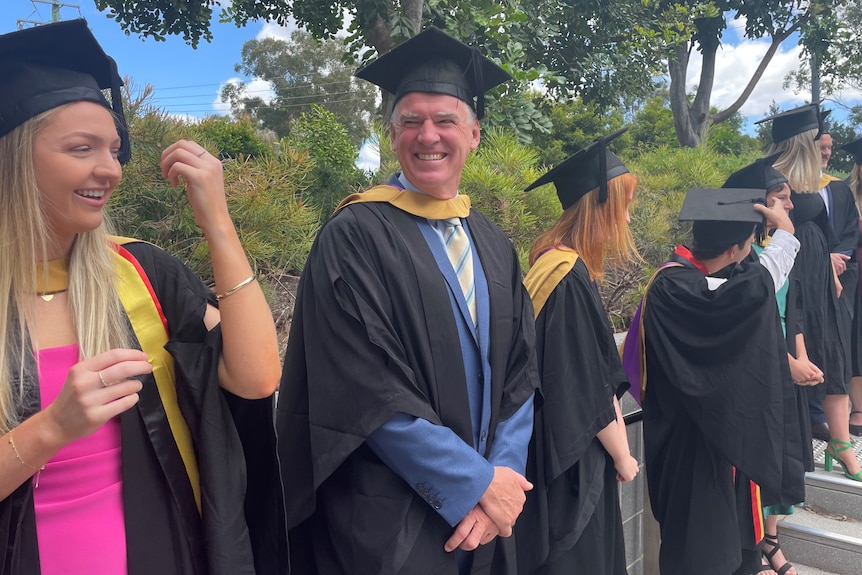 A man stands in a university graduation robe, smiling.