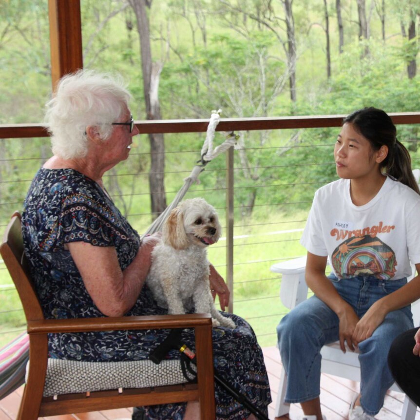 An older woman sits with two younger women on a balcony. A small white dog is on her lap.