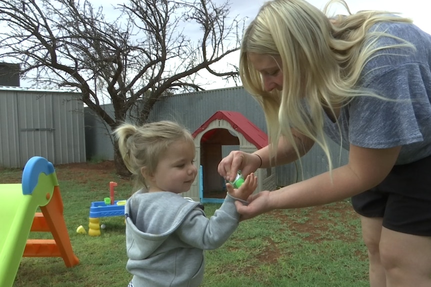 A woman with long blonde hair squirts hand sanitiser onto a young girl's hands in front of bright outdoor toys.