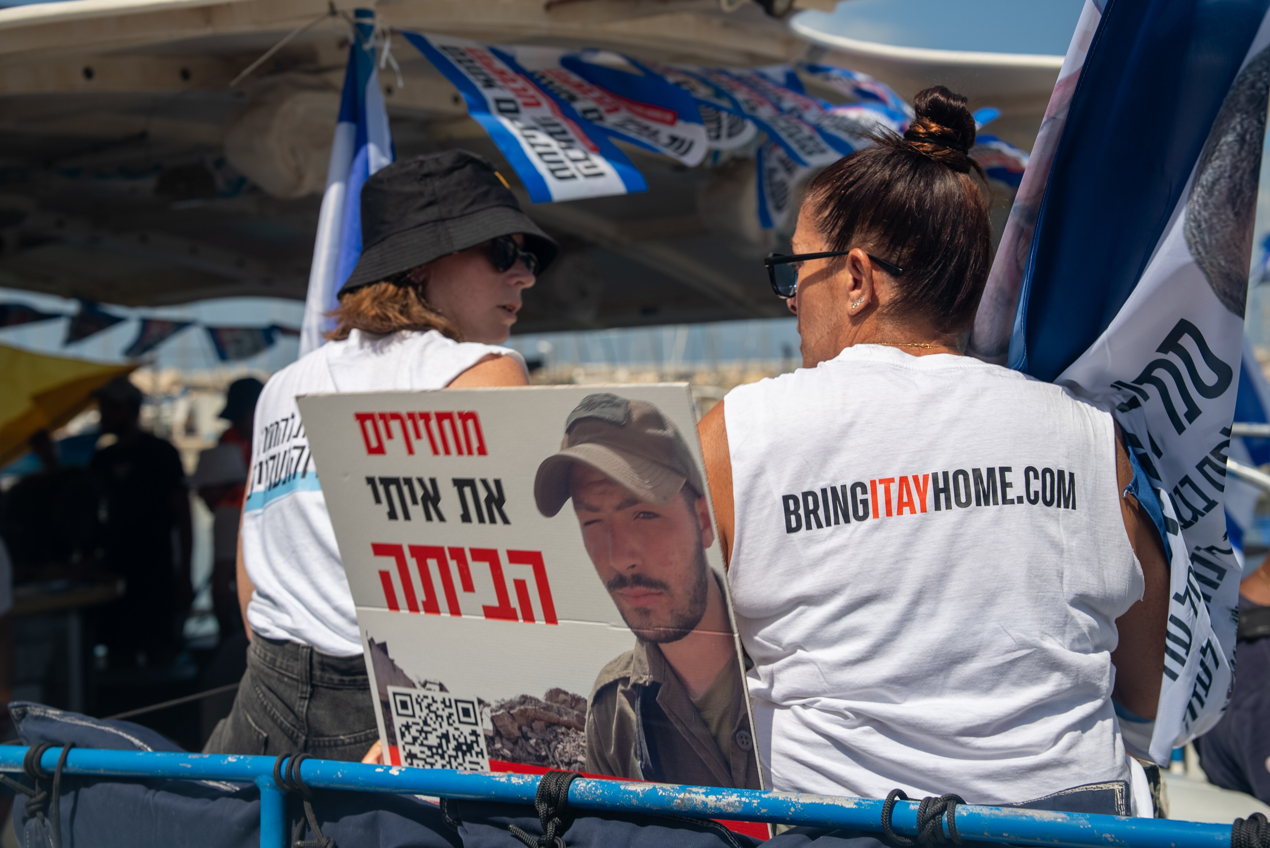 A photo of two women sitting on a boat. Their shirts say "bring them home". A placard of a hostage is held up.