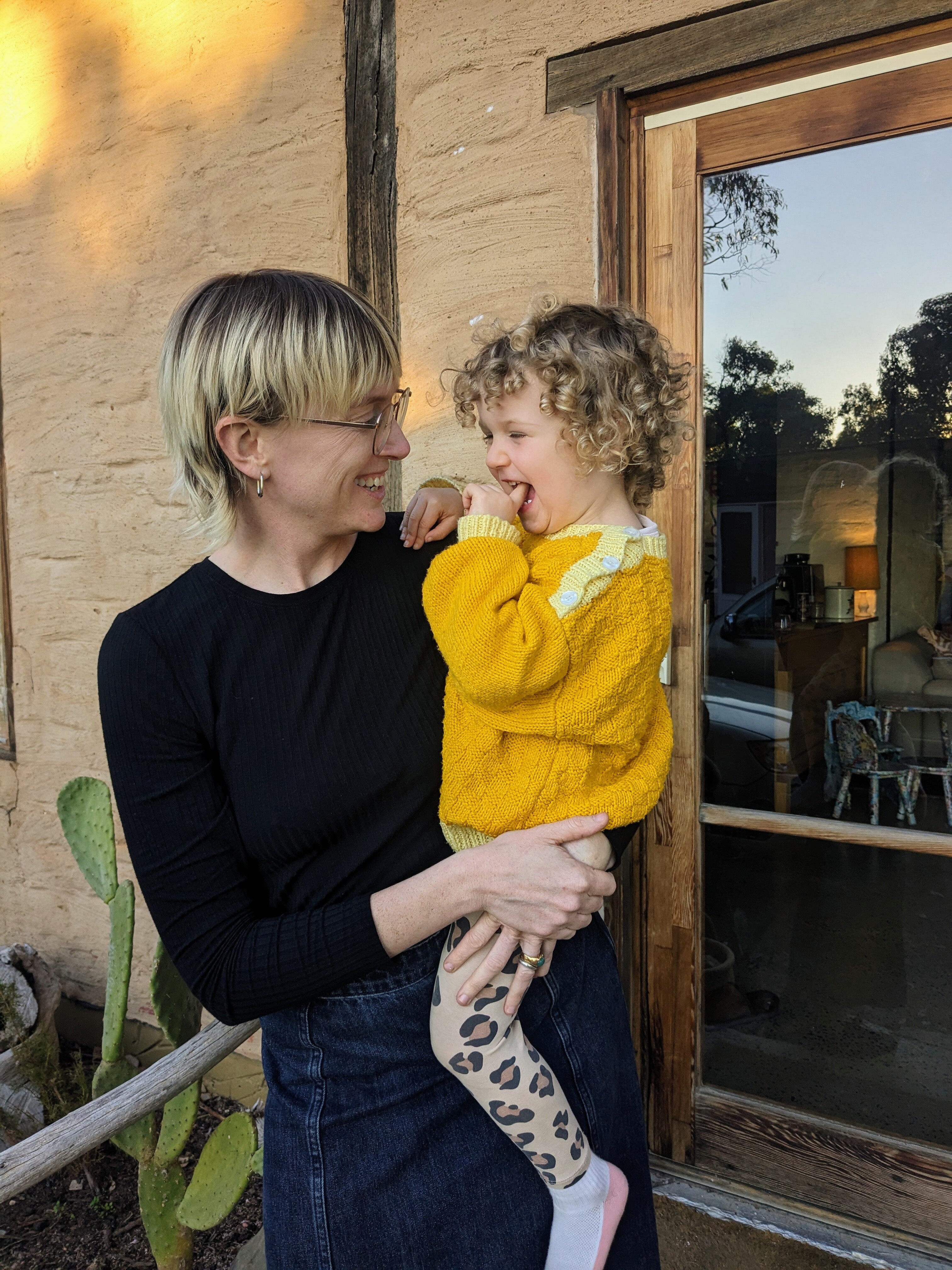 A young woman smiles with a toddler in yellow jumper sitting on her lap
