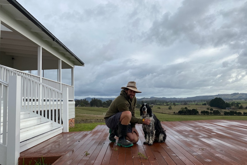a farmer kneels with his dog on the front deck of a house