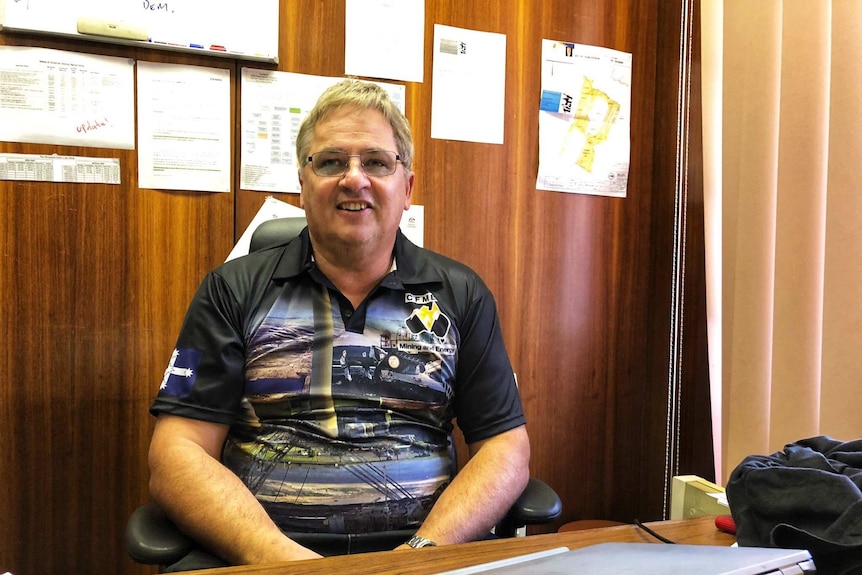 A grey-haired, bespectacled man in a dark t-shirt sits in a wood-panelled office.