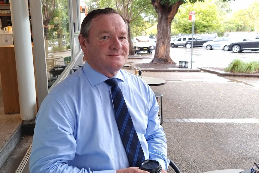 A man wearing a blue shirt and tie sits outside a cafe.