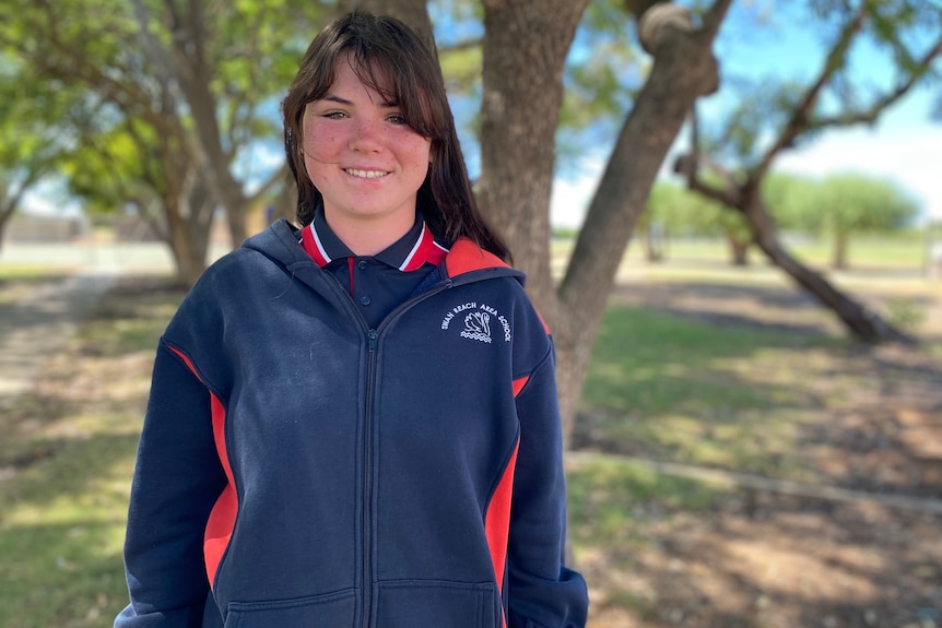 A teenager wears a navy and red school jumper, she smiles and has long brown hair with a fringe