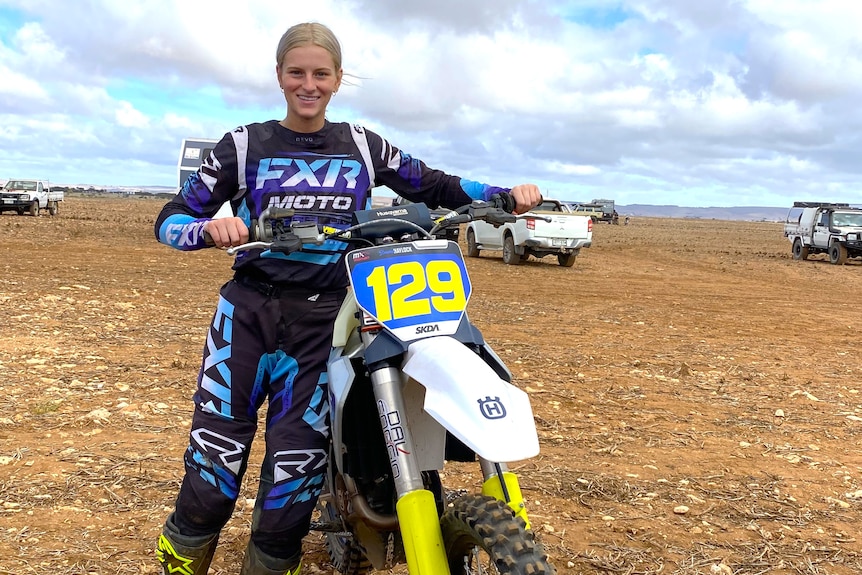 Blonde woman in riding clothes on dirt road standing with motorbike 129 looking at camera, front view of bike