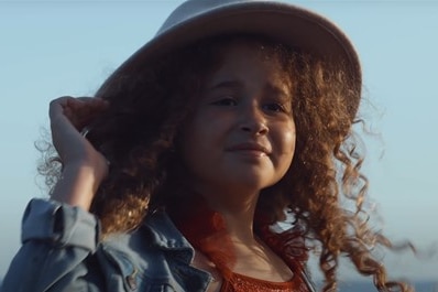 Young girl with curly hair looks at camera holding her hat. 
