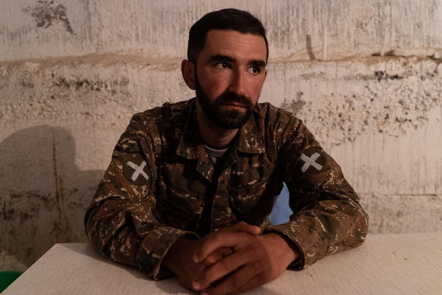 A men in a rudimentary soldier's uniform sits at a tale with his hands clasped