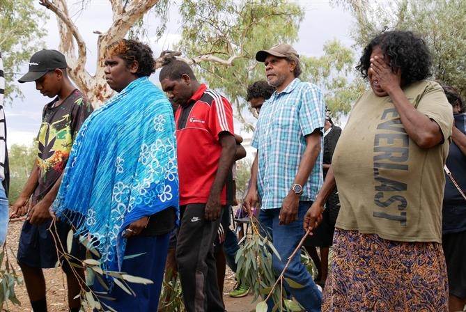 A group of Indigenous men and women carry eucalyptus leaves, head bowed, trees behind them.